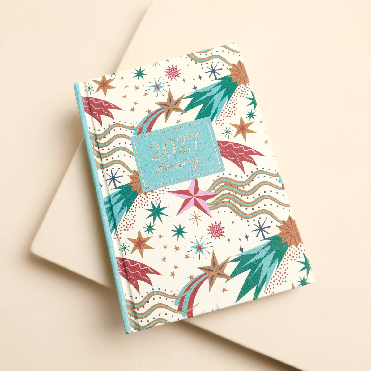 A 2027 daily planner with a colorful celestial pattern, featuring stars, planets, and waves on a cream background, styled on a neutral beige surface.