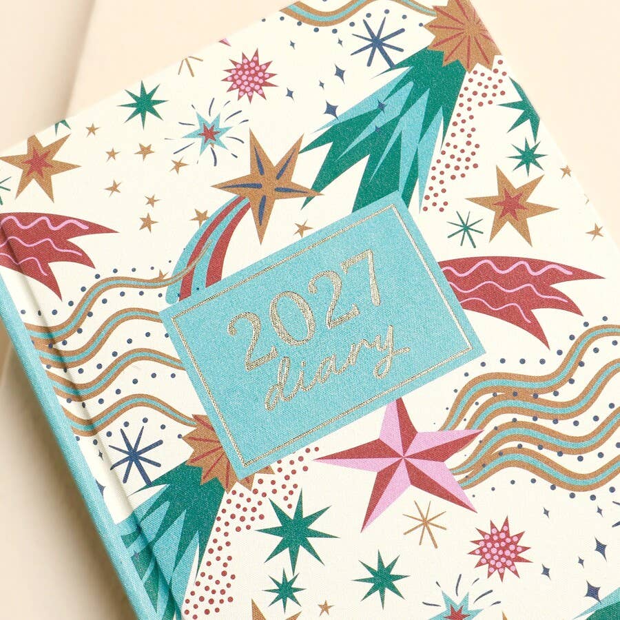 A close-up of a 2027 diary with a colorful, festive cover design featuring fireworks, stars, and wavy lines on an off-white background, with a light blue label reading '2027 diary'.