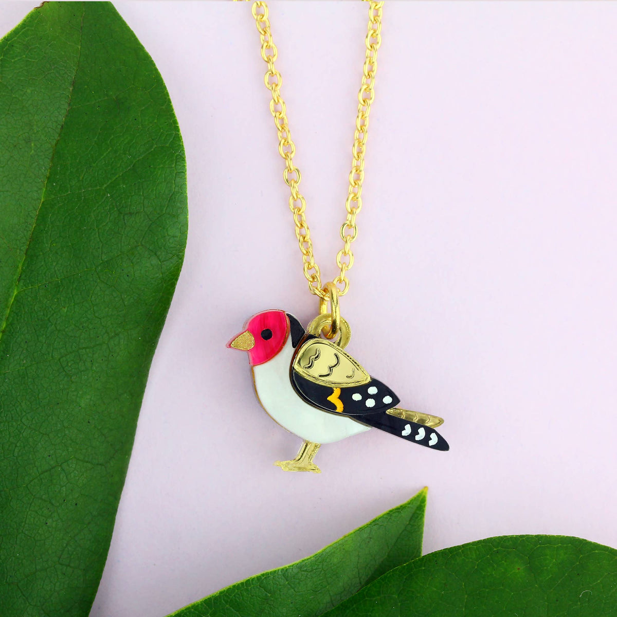 A gold chain necklace with a bird-shaped pendant, styled with green leaves on a light pink background. The pendant features a white body, a pink head, black and gold wings, and white spots on its tail.