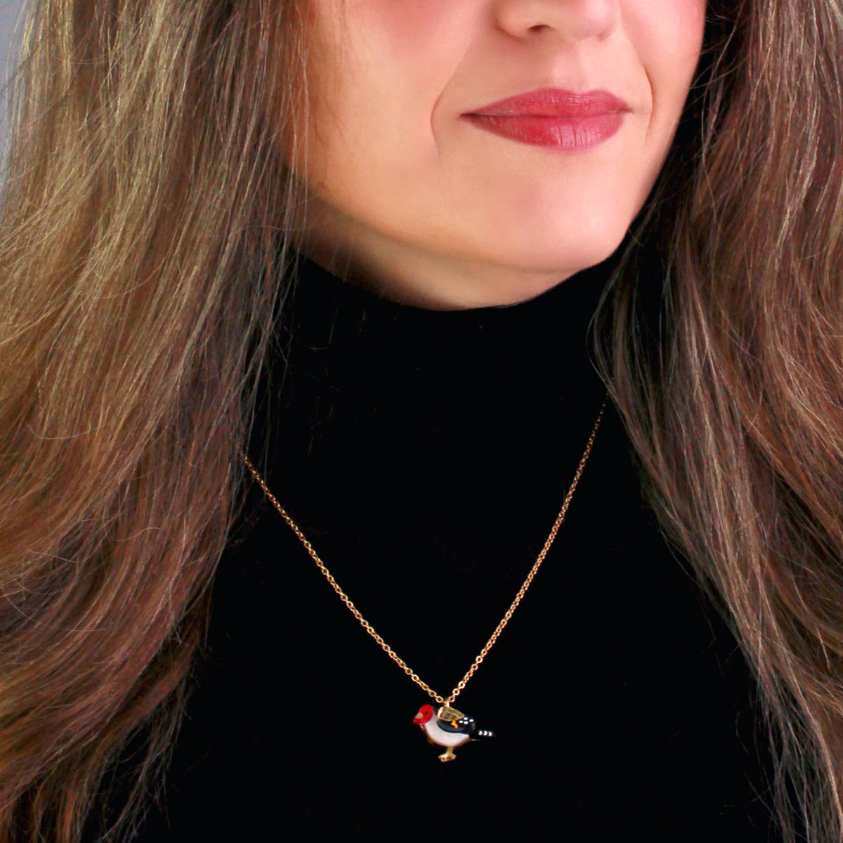 A close-up of a woman wearing a delicate gold chain necklace with a small bird pendant. The pendant features a red crest and white underbelly.