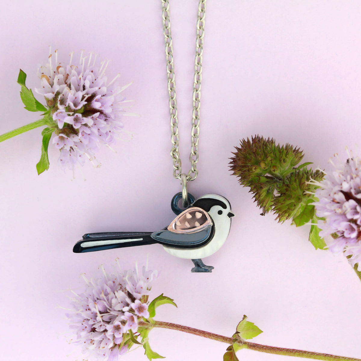 A bird-shaped pendant necklace on a silver chain, styled with dried flowers on a light pink background. The pendant features a black and white bird with a metallic rose gold wing.