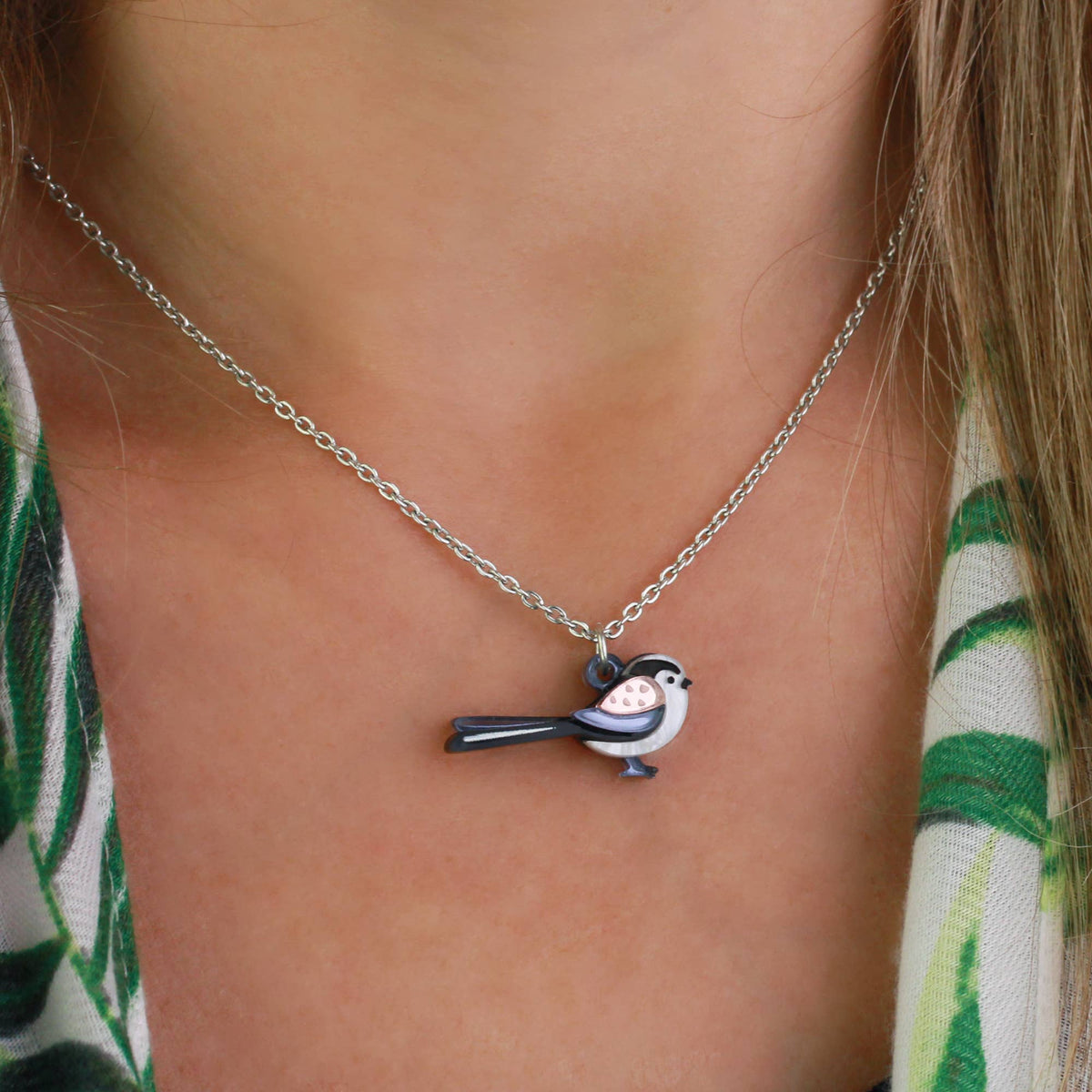 A close-up of a person wearing a delicate silver chain necklace with a small bird charm. The bird charm has black and white enamel detailing with a pink belly accent.