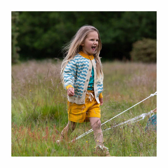 A little girl runs through a grassy field wearing a striped cardigan and yellow shorts.