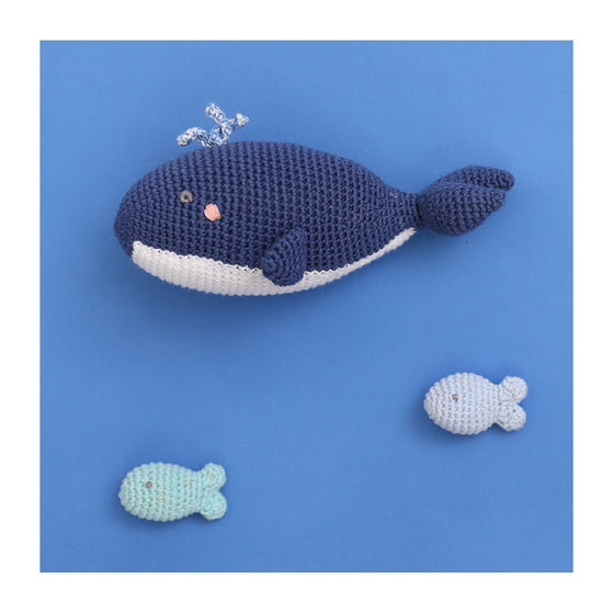A crocheted baby rattle in the shape of a whale. Wilbert is navy blue with a white under belly, pink cheeks and blowhole detailing.